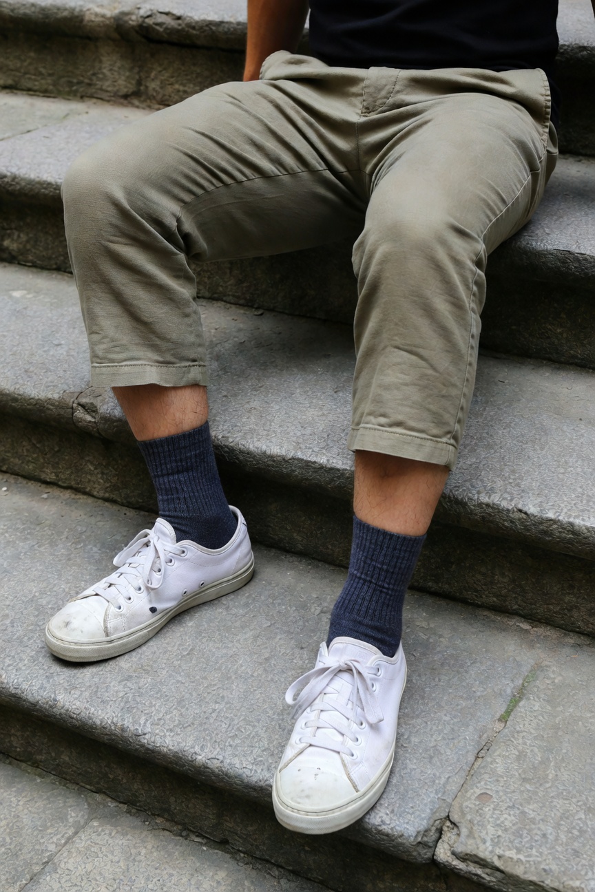 A man sits casually on stone outdoor stairs in a black t-shirt, light khaki cropped pants cuffed at the ankles, navy crew socks, and white laced sneakers, evoking relaxed summer urban style
