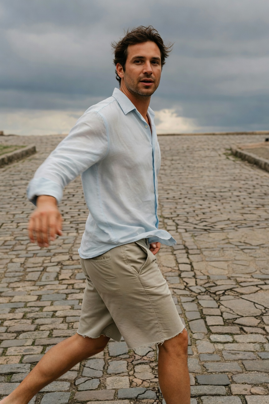 Man in his 30s with tousled dark hair walks barefoot on cobblestone path by stormy sea, wearing open light blue sheer long-sleeve linen shirt and beige tailored mid-thigh shorts, evoking elevated European summer casual style