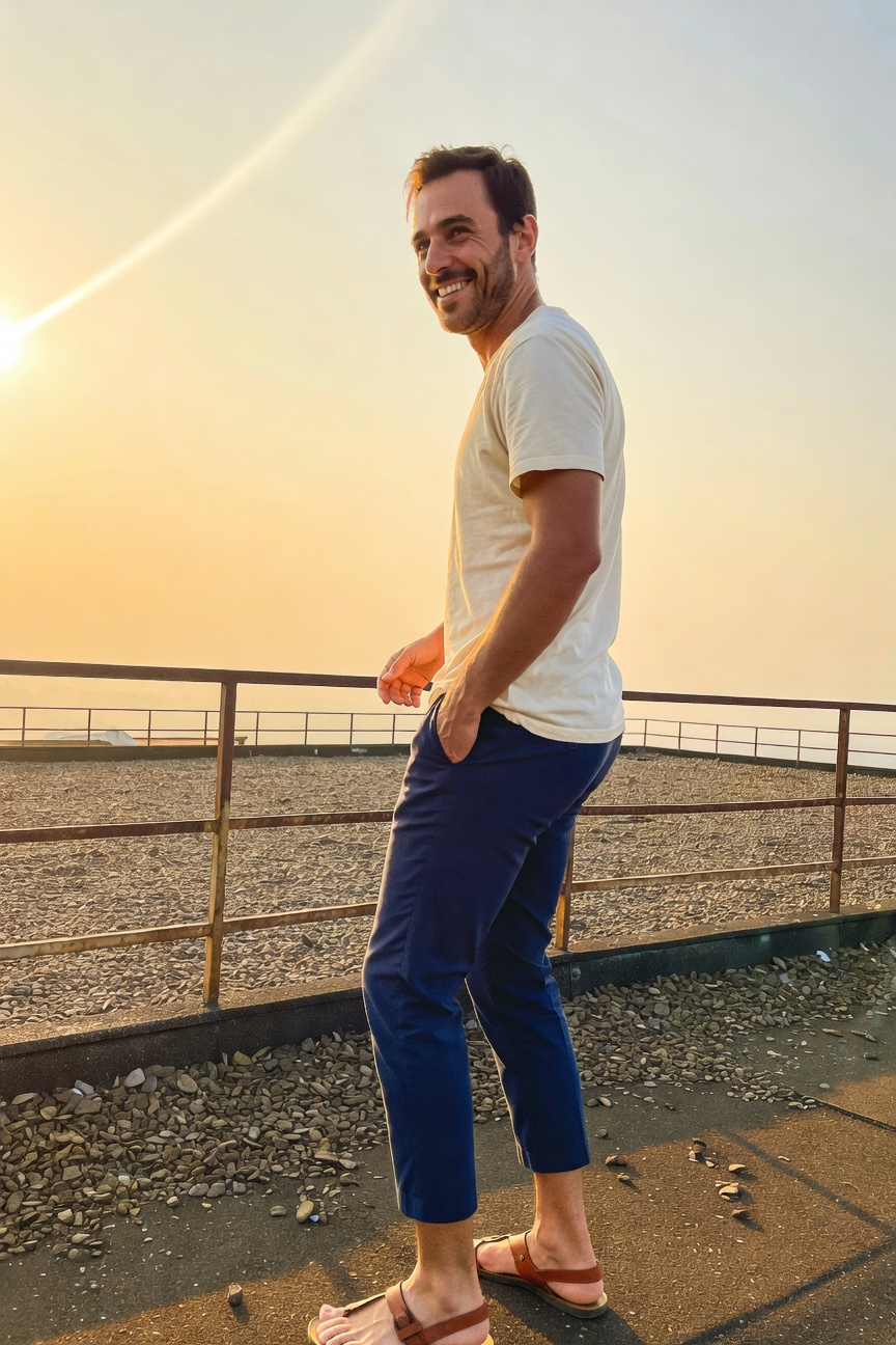 Side profile of a smiling man in a white short-sleeve t-shirt, slim navy cropped pants, and brown strap sandals, leaning on a rooftop railing at sunset with warm orange sky and hazy horizon